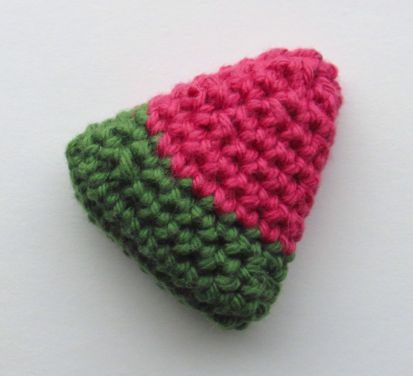 a closeup photo of a crochet piece of watermelon. It is triangular in shape with the bottom third in green and the top two thirds in pink.