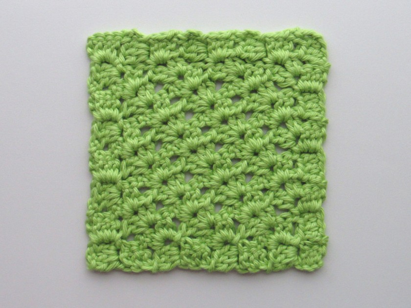 A photo of a green C2C crochet square made up of rows of V-stitch shell stitches with a border of sets of 3 dc stitches around the square. The V-stitch shell stitch is a set of four dc stitches with a chain stitch in the middle.