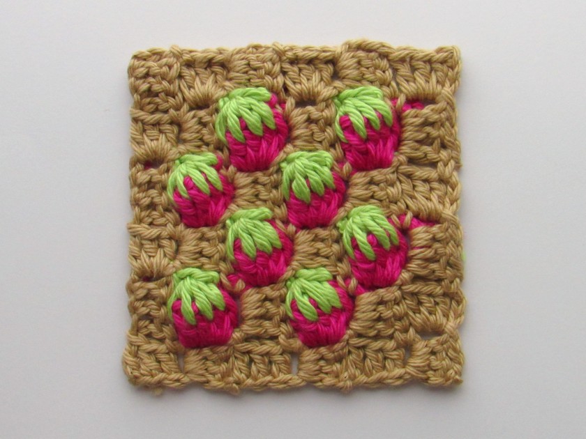 a photo of a c2c crochet strawberry stitch square. the non-strawberry stitch parts are a light brown color, and the strawberries are red and green.